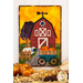 A photo of an autumn-themed wall hanging featuring a red barn, wagon full of pumpkins, and a sheep hanging on a white wall with fall decor of straw, dried corn, and small pumpkins in the foreground.