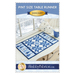 Front cover of the pattern featuring title, designer and photo of finished project--A blue and white winter-themed table runner atop a white counter with a wintry scene outside an adjacent window and snowflake decorations with a mug of hot cocoa