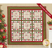 Photo of a Christmas themed quilt featuring 9 blocks with embroidered motifs hanging on a cream colored wall with a Christmas tree on one side and holiday decor on the other.