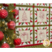 Close up photo of a Christmas themed quilt featuring 9 blocks with embroidered motifs hanging on a cream colored wall with a Christmas tree in the foreground