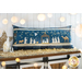 Photo of a large blue bench pillow embellished with decorative stitching in a nativity scene with twinkling lights atop a farmhouse style cabinet with hay, lights, and small nativity decor all around with a white paneled wall in the background
