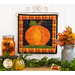 A square wall hanging featuring a pumpkin on a craft holder hanging on a white paneled wall with autumn decor in the foreground.