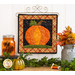 A square wall hanging featuring a pumpkin on a craft holder hanging on a white paneled wall with autumn decor in the foreground.