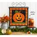 A square wall hanging featuring a jack-o-lantern on a craft holder hanging on a white paneled wall with autumn decor in the foreground.