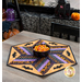 Photo of a Halloween themed table topper in the center of a gray table with halloween decor in the background and a bowl of candy corn in the center of the table.
