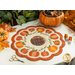 Photo of a scalloped table mat with pumpkins in a ring and embroidered details on a white table with a pair of scissors, threads, and pumpkin decor
