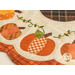 Close up photo of a scalloped table mat with pumpkins in a ring and embroidered details around a brown plaid circle at the center