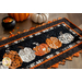 Close up photo that shows fabric detail and stitching of a Halloween themed table runner made with black, orange, and white fabrics with a row of pumpkins in the middle, laying on a wooden table top with pumpkins and a small decorative cauldron filled with candy corn.