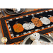 Close up photo of a Halloween themed table runner made with black, orange, and white fabrics with points along the top and bottom and a row of pumpkins in the center, on a wooden table top with Halloween decor