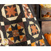 Close up photo of an orange, brown, and black quilt featuring traditional quilt block patterns draped in front of a brown paneled wall with fall decor in the background