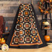 Photo of an orange, brown, and black quilt featuring traditional quilt block patterns draped in front of a brown paneled wall with fall decor all around