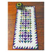 Photograph of the finished vortex table runner, including a white background with purple and green triangles on it, set on a wooden table 