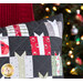 Close up photo of a chair with a red blanket draped over it with a decorated christmas tree in the background and a pillow featuring small quilted gifts motifs all over.
