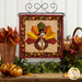 Photo of the finished foundation paper piecing block for November featuring a turkey dressed in a tie and capotain hanging from a tabletop craft holder and surrounded by fall decor like leaves and pumpkins