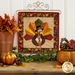 Photo of the finished foundation paper piecing block for November featuring a turkey dressed in a tie and capotain hanging from a wall mounted craft holder and surrounded by fall decor like leaves and pumpkins