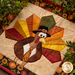 Close up photo of the finished paper piecing block showing stitching and fabric details of a cartoon turkey wearing a tie and capotain, surrounded on a countertop by foliage