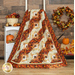 Photo of the Reflections of Autumn Log Cabin Throw Quilt draped over a white ladder in front of a dark paneled wall with a small white shelf to one side, an autumn wreath, and pumpkin decor
