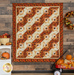 Photo of the Reflections of Autumn Log Cabin Throw Quilt hanging flat on a dark paneled wall with a small white shelf to one side, an autumn wreath, and pumpkin decor