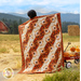 Photo of the log cabin throw quilt made with orange, brown, and cream fabrics held across the shoulders of a woman with a red wagon filled with pumpkins in a field with mountains and a red barn in the background