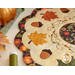 Close up photo of an autumn-themed table topper on a white table with fall decor all around including leaves, a gold pumpkin, and thread spool