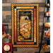 Photo of a Thanksgiving and autumn-themed panel quilt hanging on a brown paneled wall with fall decor like pumpkins, candles, and books on a shelf with a floral basket on a chair in the foreground