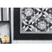 Close up photo of one corner of a black, white, and gray geometric quilt with floral patterns in the fabrics, hanging on a white wall next to a black shelf with matching, monochrome decor.