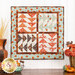 Photo of a wall hanging designed in quadrants of flying geese units made with aqua, orange, cream, and brown autumn themed fabrics hanging on a white paneled wall with fall decor on the countertop below it
