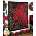 An angled shot of the finished Poinsettia Kaleidoscope quilt hung flat on a white-paneled wall with a brown tiered shelf holding coordinating decor and a black chair draped in a red blanket with a vase of poinsettias sitting on it. Above the chair hangs a poinsettia wreath and along the floor below the quilt, a thick garland of poinsettias, pinecones, and christmas tree branches decorates the width of the shot.