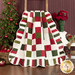 Photo of a green, red, and cream Christmas-themed quilt draped over a ladder in front of a red wall with a Christmas tree and poinsettia decor in the background.