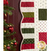 Close up photo of a green, red, and cream Christmas-themed quilt flat against a wall with a decorated Christmas tree in the foreground