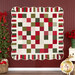 Photo of a green, red, and cream Christmas-themed quilt hanging on a red paneled wall with a Christmas tree on one side and poinsettia decor on the other