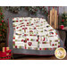 Photo of the Hometown Christmas quilt draped over some furniture in front of a gray wall with Christmas decor all around including a wooden sled, Christmas tree, wrapped gifts, and pine foliage.