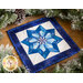 Photo of the completed quilt block featuring a pinwheel shaped snowflake made with different shades of blue fabric with a white background and dark blue border laying flat atop a wooden counter with winter garland all around.
