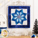 Photo of the completed quilt block featuring a pinwheel shaped snowflake made with different shades of blue fabric with a white background and dark blue border hanging on a wall hanging craft holder above a wooden counter with winter decor.