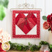 Photo of the completed February project featuring a red heart and key charm on a white background with a red border hanging on a craft holder on a white paneled wall with floral decor on either side.