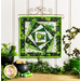 Photo of the finished wall hanging featuring offset squares in shades of green with a shamrock and top hat in the center, on a craft holder on a white paneled wall and decorative foliage.