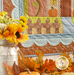 A view of the bottom left corner of the quilt from an angle, demonstrating fabric and top quilting details. A blurry milk can full of sunflowers and scattered leaves on the floor are in the foreground of the shot.