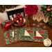 Photo of four Magic Stained Glass Placemats arranged in a fan on a wooden table with a set of 4 wine glasses, plates, and matching cloth napkins with a small wrapped gift, decor, and poinsettias in the background.