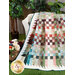Photo of a quilt made with natural colors draped over a small white bench with a basket of flowers in the grass and plants in the background.