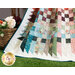 Close up photo of a quilt made with natural colors draped over a small white bench with a basket of flowers in the grass