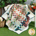Photo of a quilt made with natural colors draped over a small white bench with a basket of flowers in the grass on one side and a vase of flowers sitting on the bench on the other