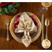 Photo of a cream cloth napkin in a napkin ring on a plate edged with gold, with gold silverware, a wine glass, and foliage on a wooden table