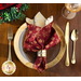 Photo of a red cloth napkin in a napkin ring on a plate edged with gold, with gold silverware, a wine glass, and foliage on a wooden table