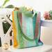 Photo of a large jelly roll tote bag filled with flowers sitting on a small white chair with a white table, houseplant, and bouquet in the background.