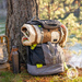 A camping backpack with the Paddling Bears quilt, rolled up and tucked into the backpack with a bear face peeking out, leaning against a tree with a river in the background.