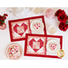 A top-down angle of the two completed Tea & Cookies for Two - February placemats, staged on a white table with red roses, white teacups, a pearlescent pink teapot, and frosted heart shaped cookies with pink and red sprinkles.