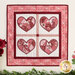 Photo of a pink, red, and white wall hanging featuring four red hearts in quadrants, hanging on a white paneled wall with foliage on the countertop below.