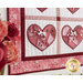 Close up photo of a pink, red, and white wall hanging featuring four red hearts in quadrants, hanging on a white paneled wall with a bouquet of roses in the foreground.
