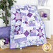 Photo of a purple, blue, and white quilt made with a psalms panel draped on a chair in front of a white paneled wall with white furniture and purple floral decor on either side.