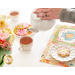 A model pours a cup of tea from a white teapot; the placemat and plates of cookies are in view.